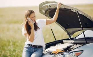 Title image for UM and UIM Coverage, Exclusions, and Endorsements showing a woman stranded in the middle of the road sitting on the hood of her car calling her insurance carrier.